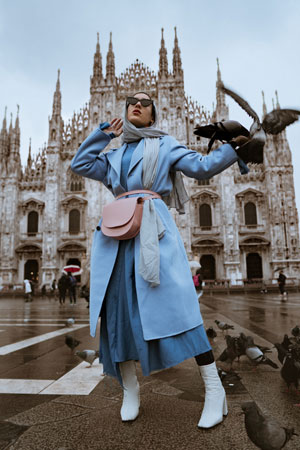 A photographic image of a girl wearing sunglasses, a blue dress with a blue long coat, and a blue headscarf. She is looking up from the camera, with her arm outstretched for birds to perch on.