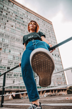 A photographic image of a girl, leaning on a handrail in front of a building. She is looking down at the camera with her foot posed before stepping on the camera lens.
