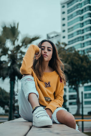 A photographic image of a girl who is sitting on a bench in front of palm trees and a skyscraper, and poses while looking at the camera.