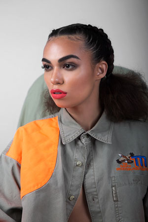 A photographic image of a girl with bright lipstick, wearing a jacket with an orange shoulder patch as she looks away from the camera.