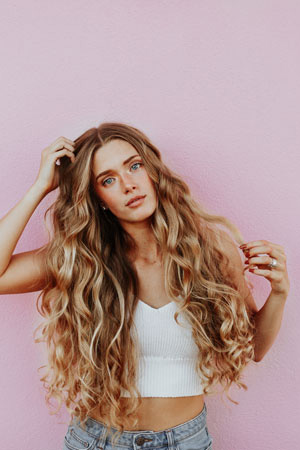 A cropped photographic image of a girl with long, blonde, and wavy hair posing in front of a pink wall as she looks at the camera.