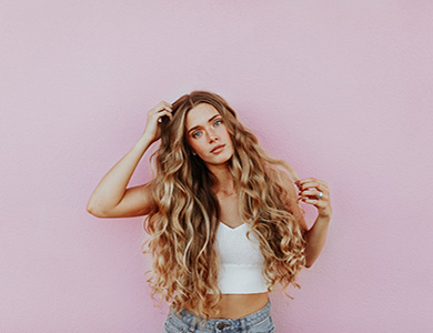 A photographic image of a girl with long, blonde, and wavy hair posing in front of a pink wall as she looks at the camera.