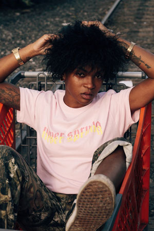 A photographic image of a girl sitting in a shopping cart. She looks at the camera lens as her hands rest in her curly hair.