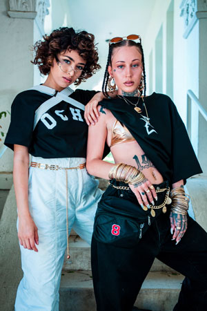 A photographic image of two girls wearing several gold accessories, both looking at the camera as they pose closely together in front of building steps.