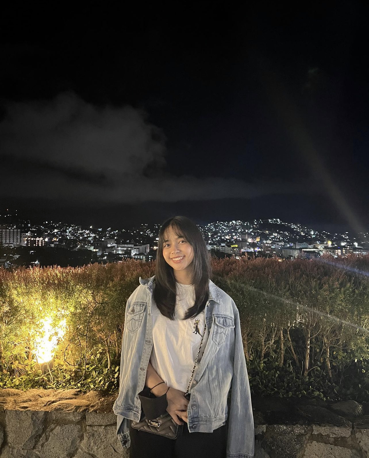 A picture of girl smiling with the background being an overlook of a city at night.