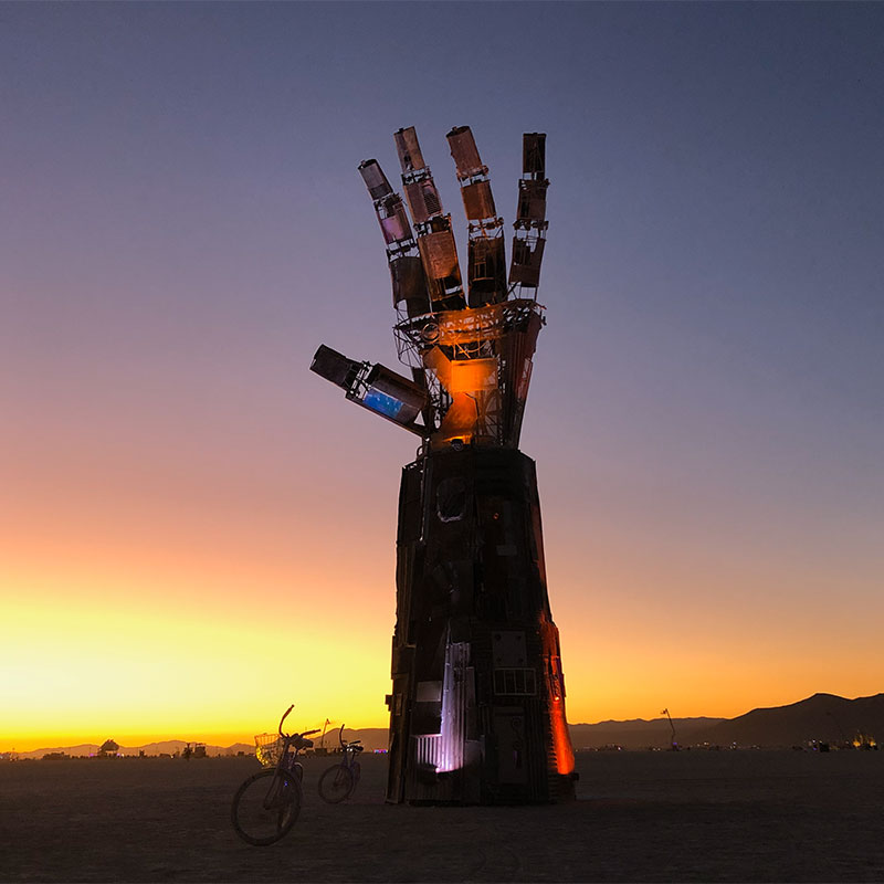 A giant mechanical hand sculpture lit up with red and purple lights sits in the desert with a purple and yellow sky just after sunset