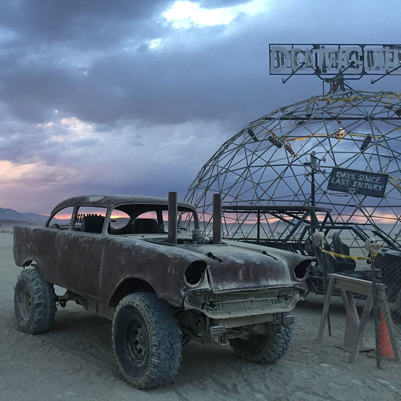 A 1950's car with lifted wheels sits next to a metal dome with a purple and grey cloudy sky