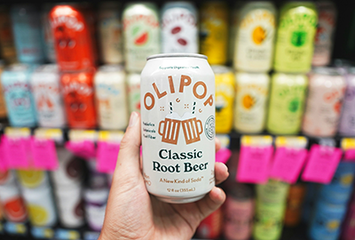 Hand holding Olipop root beer soda in front of a colorful drink shelf