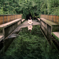 A woman sits at the end of a bridge with a reflecting pool of water in front of her.