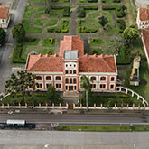 Tan school building with terracotta roofing next to a large lawn filled with hedges of bushes, pathways, and small trees. 