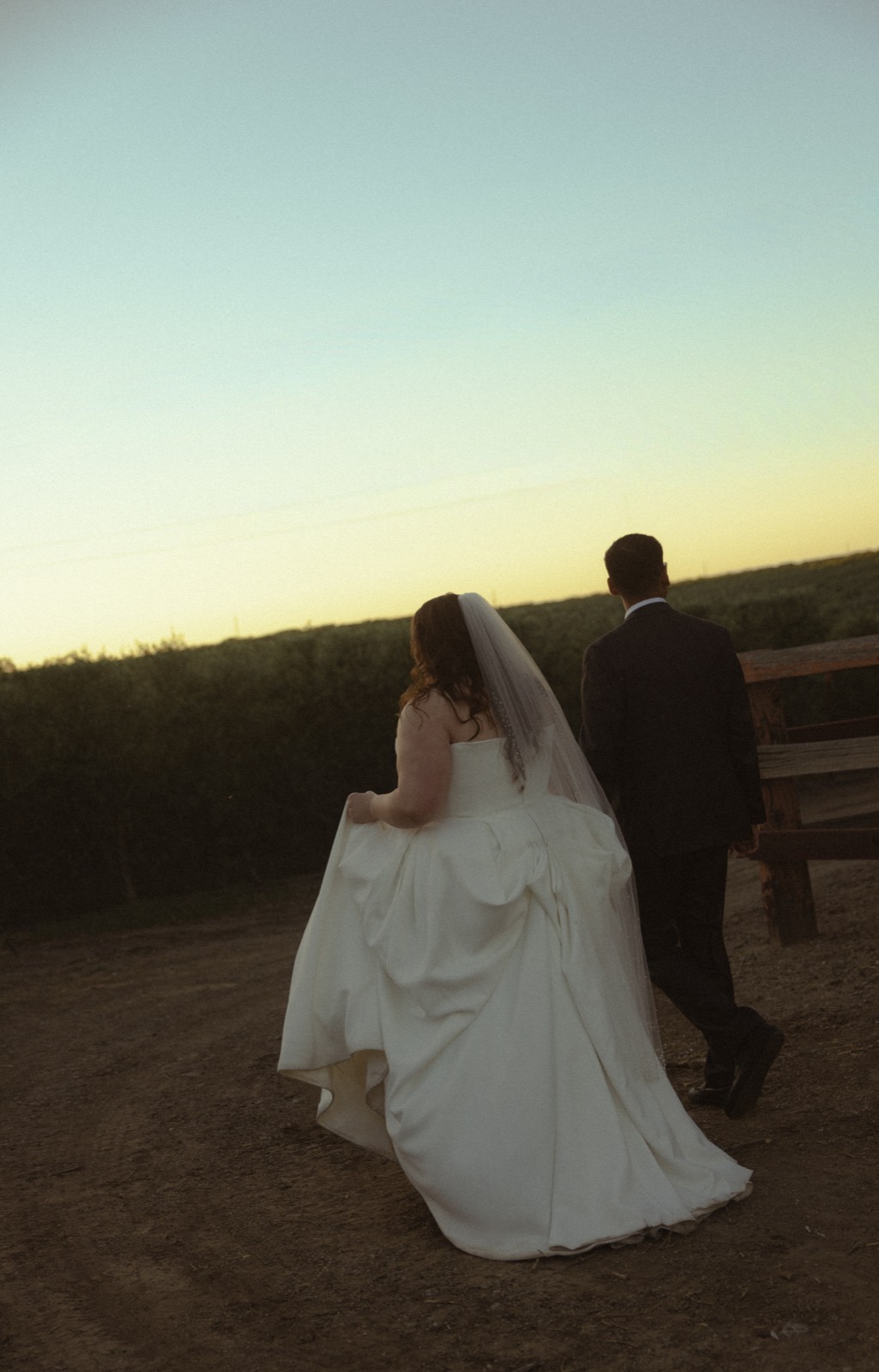 A bride and groom walking together at sunset along a dirt path lined with hedges.