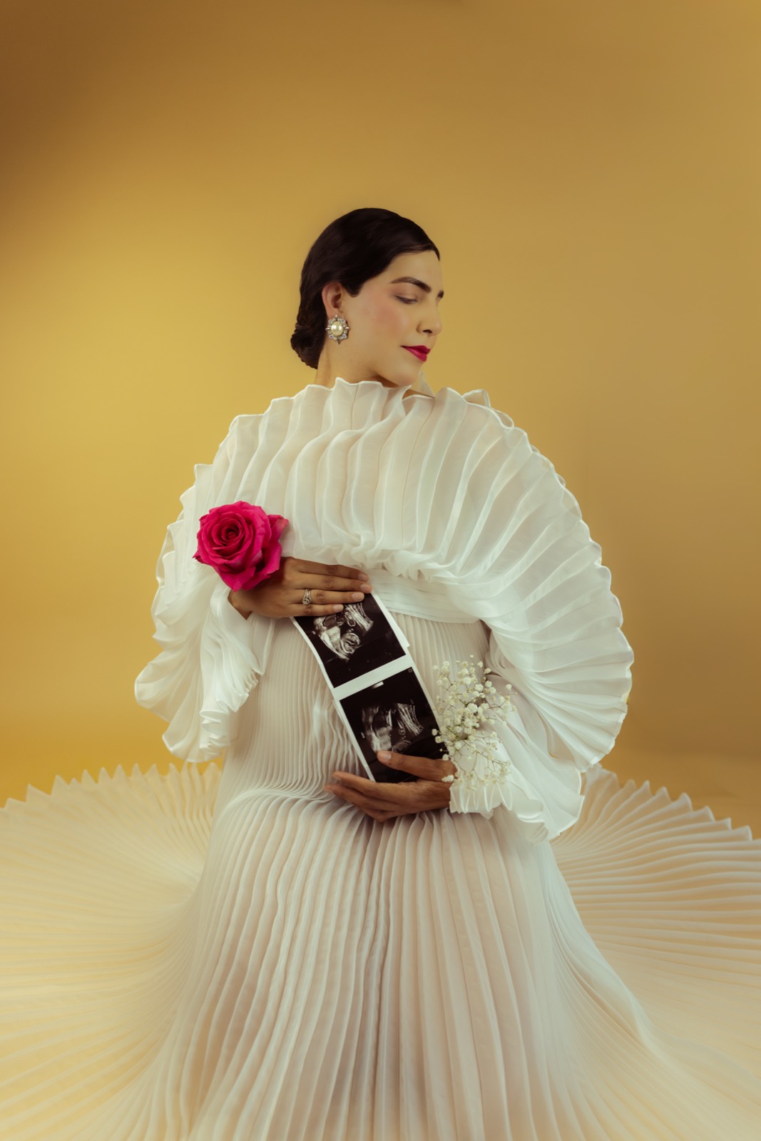 An editorial portrait of a woman in a pleated white gown holding ultrasound photos and a pink rose.
