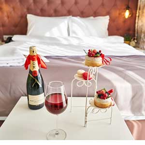 a bed with fresh towels and a small table in front with a wine bottle, a glass of wine and little cakes