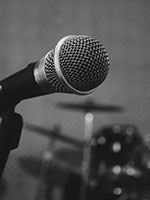 Closeup greyscale picture of a microphone on a stand and a drumset behind it.