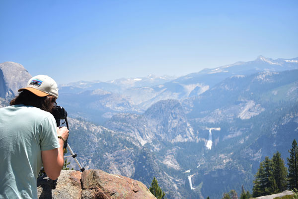 College age man with long hair at the corner of the photo looking through the view finder of a camera. Half Dome and the waterfalls near it at the close of the Yosemite Valley in the background.